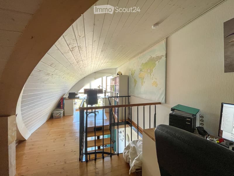 Office space with wooden flooring, slanted ceiling, and a railing overlooking a lower level. Includes a desk, chair, laptop, bookshelf, and a world map on the wall.