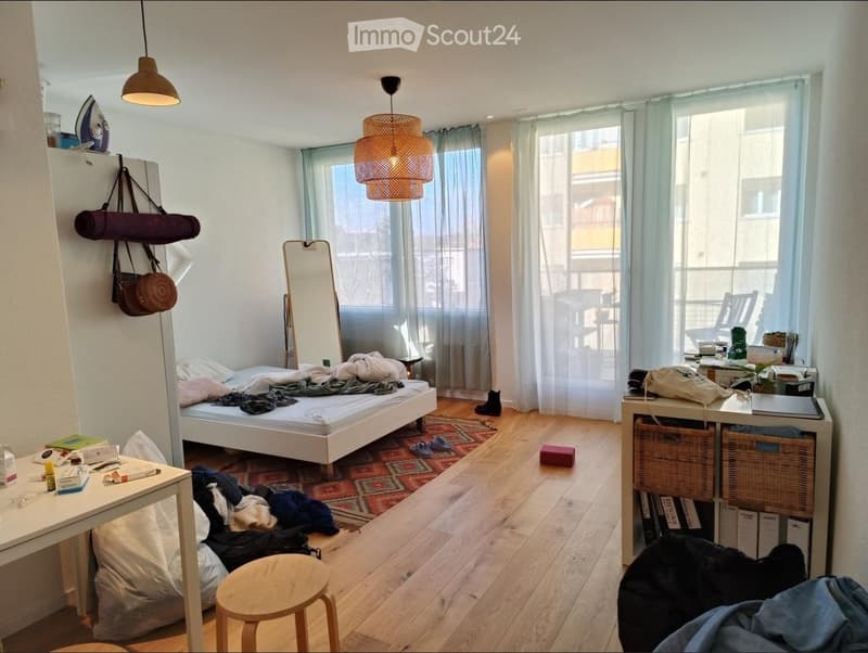 A small, well-lit bedroom with a bed, a mirror, a small table, a wicker shelf, and a hanging pendant light. The room features wooden flooring and large windows with sheer curtains.