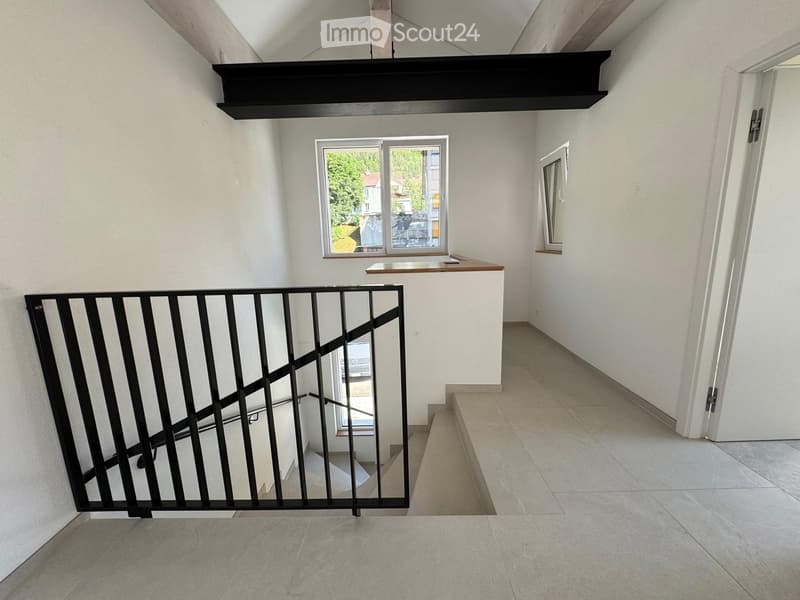 Empty hallway with tiled floor, white walls, and a window at the end. A black metal railing leads to a staircase, and a small wooden counter is visible near the window.