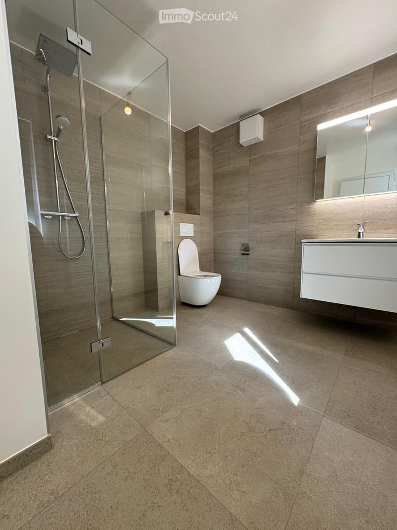Modern bathroom with beige tiled walls and floor, glass-enclosed shower, toilet, sink, and illuminated mirror.