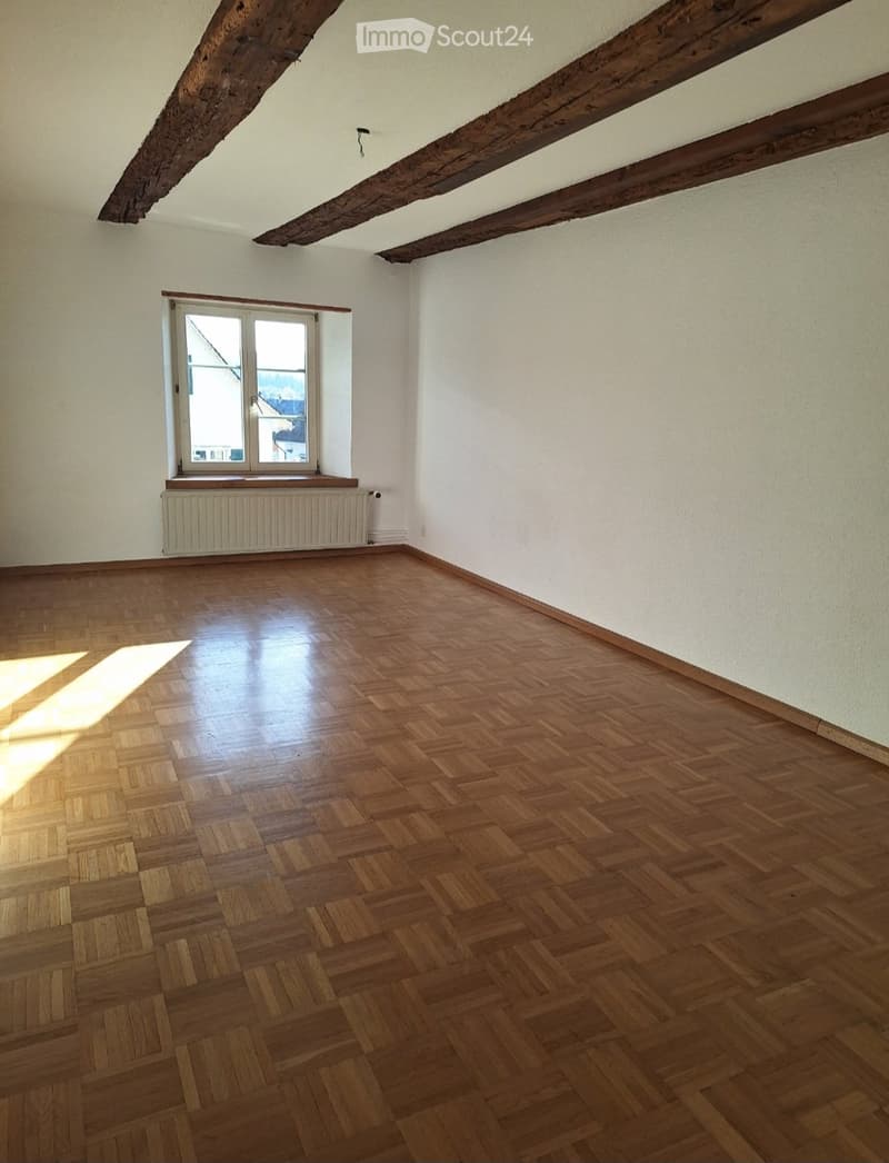 Empty room with wooden herringbone flooring, white walls, exposed wooden beams on the ceiling, and a single window with a radiator below it.