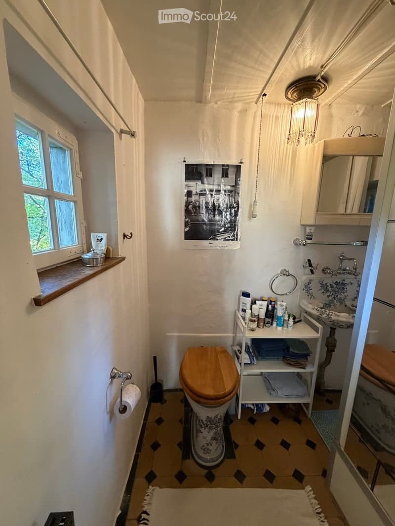 Cozy bathroom with a wooden toilet seat, sink, mirror, and shelves holding towels and toiletries. The room features a window with a view of greenery, a picture frame on the wall, and a small shelf with various items.