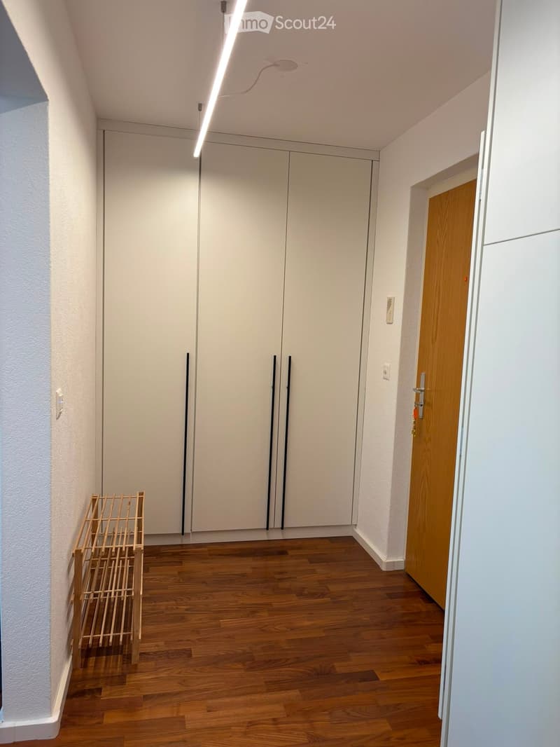 Empty hallway with wooden flooring, white walls, and a large white closet with double doors. A small wooden shelf is placed against the left wall.