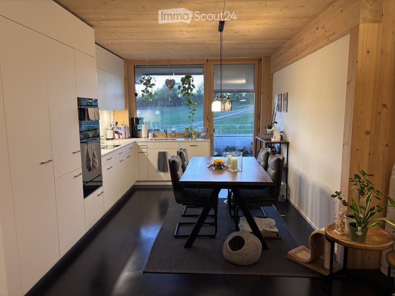 Modern kitchen and dining area with white cabinets, dark flooring, and large windows. Includes a dining table with chairs, a small side table with a potted plant, and a cat bed.