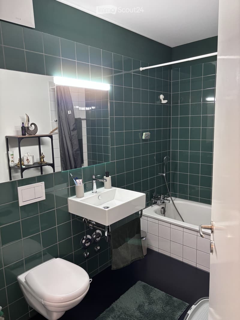 Modern bathroom with dark green tiles, white fixtures, a large mirror, a sink, a toilet, a bathtub, and a small shelf with toiletries.