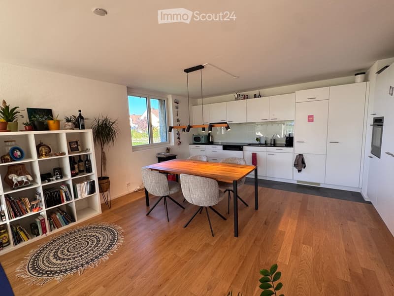 Open-plan living and dining area with a wooden dining table, four chairs, white kitchen cabinets, a microwave, and a bookshelf filled with books and decorative items.