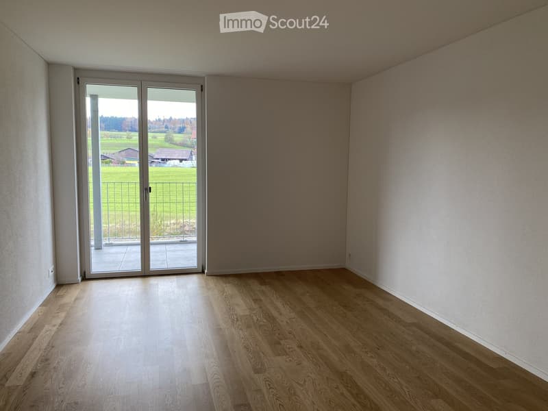 Empty room with wooden flooring, white walls, and large sliding glass doors leading to a balcony with a view of a green field.