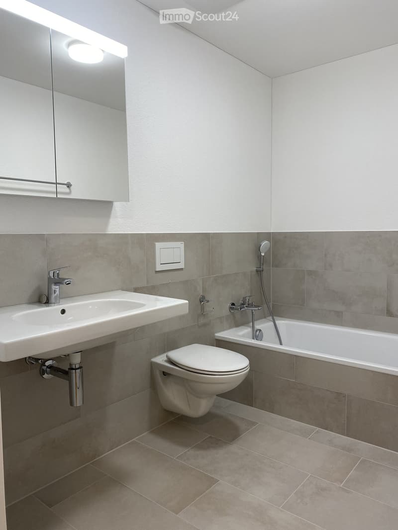 Modern bathroom with a white sink, chrome faucet, toilet, bathtub, and showerhead. Grey tiled walls and beige tiled floor.