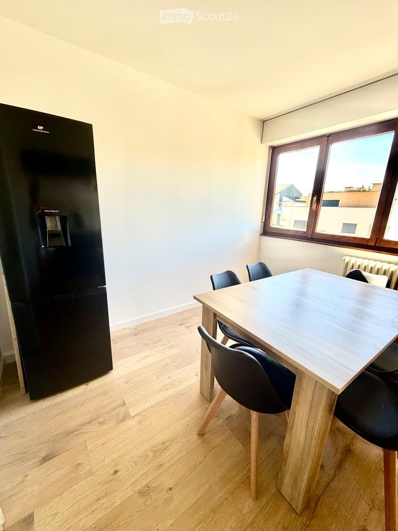 Modern kitchen with light wooden flooring, a black refrigerator, a wooden dining table, and four black chairs.