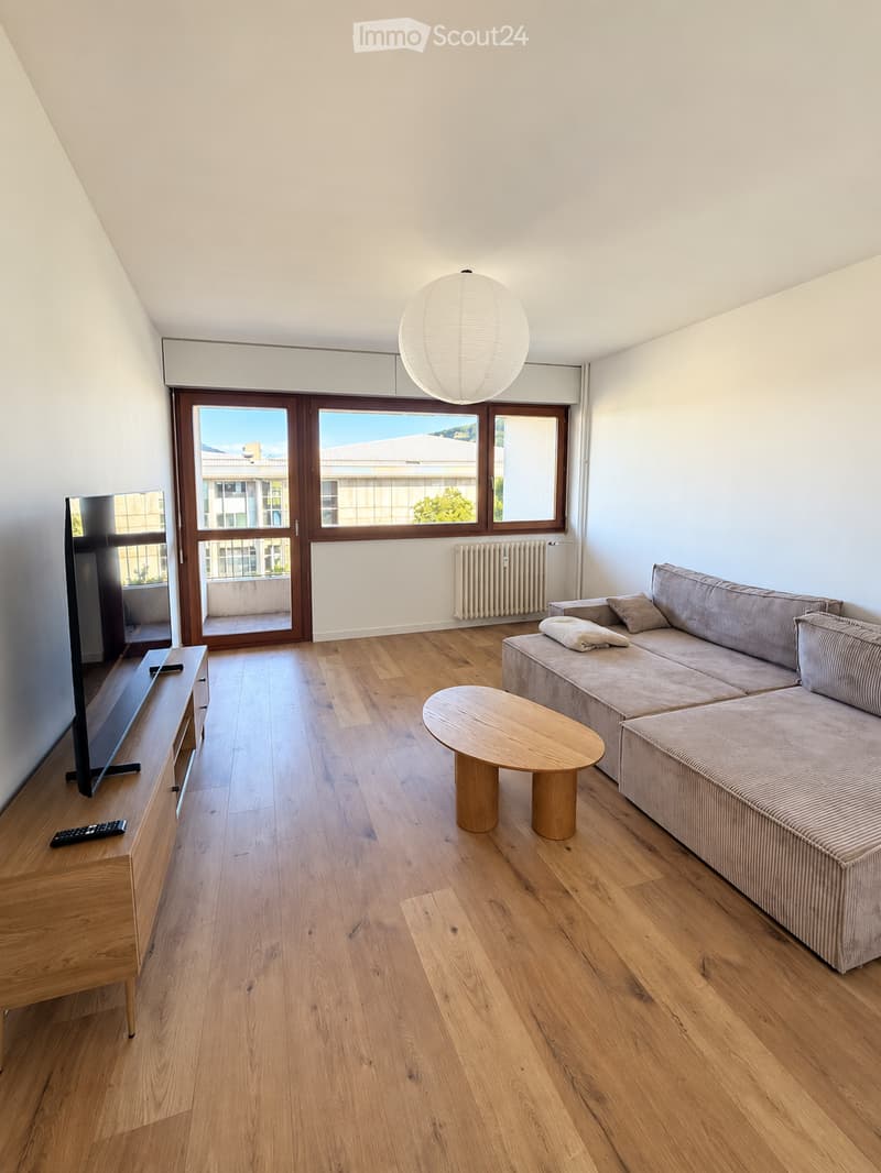 Modern living room with wooden flooring, large windows offering a view, a grey sofa, a round wooden coffee table, a flat-screen TV on a wooden stand, and a remote control on the stand.