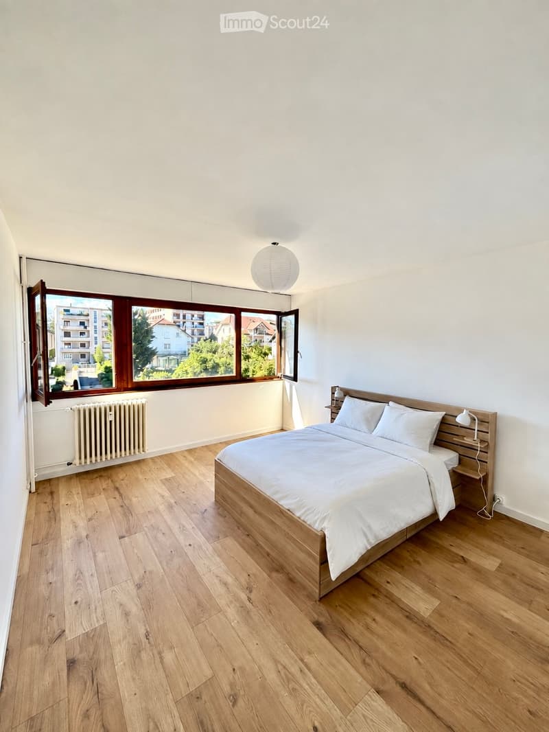 Bright bedroom with wooden flooring, large windows, a neatly made bed with white linens, and a nightstand with a lamp.