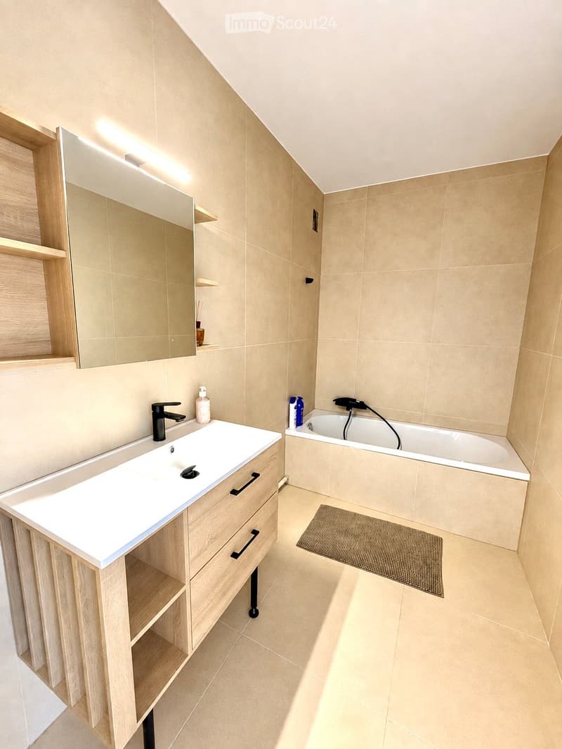 Modern bathroom with a white sink, black faucet, wooden cabinet, bathtub, mirror, and a small rug on the floor.