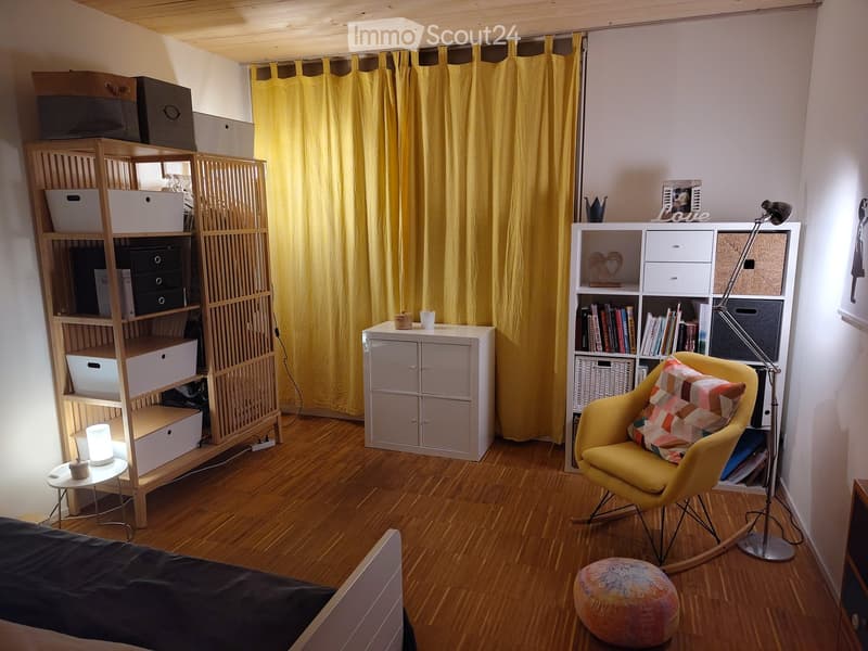 A cozy, well-furnished room with a bed, a yellow armchair, a small round table, shelves filled with books and storage boxes, and a decorative ottoman.