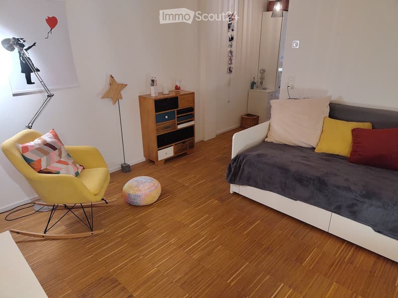 Cozy bedroom with a modern sofa, rocking chair, colorful stool, wooden cabinet, and a floor lamp. The room features wooden flooring and a playful mural on the wall.