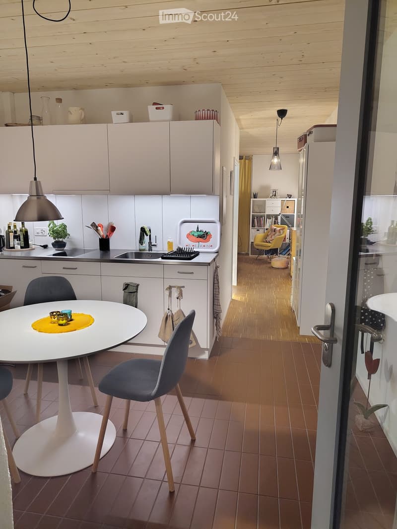 The image shows a modern, open-plan kitchen and dining area with white cabinets, a central island, and a round dining table with chairs. The kitchen features a sink, pendant lighting, and various utensils and decorative items on the counter. The dining ar