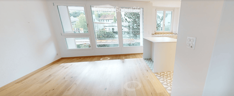 Empty room with wooden flooring, white walls, and large windows offering a view of the outside. A small kitchen counter is visible on the right side.