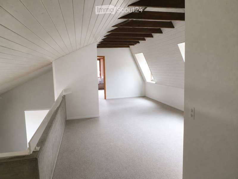 Empty attic space with white walls, carpeted floor, exposed wooden beams, and skylight windows.