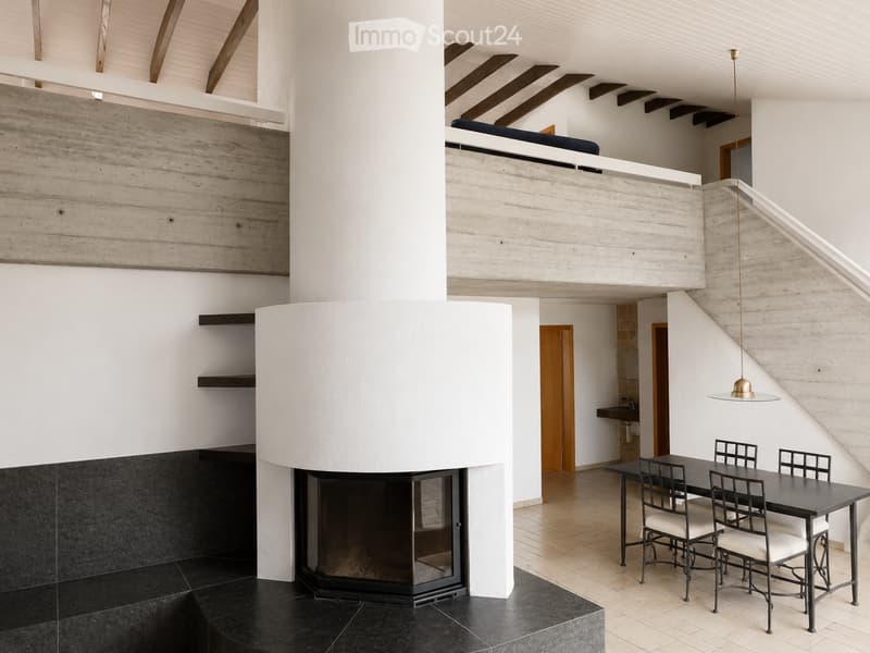 Modern open-plan living and dining area with a central fireplace, a dining table with chairs, and a hanging lamp. The space features a raised ground floor and a lofted bedroom area above.