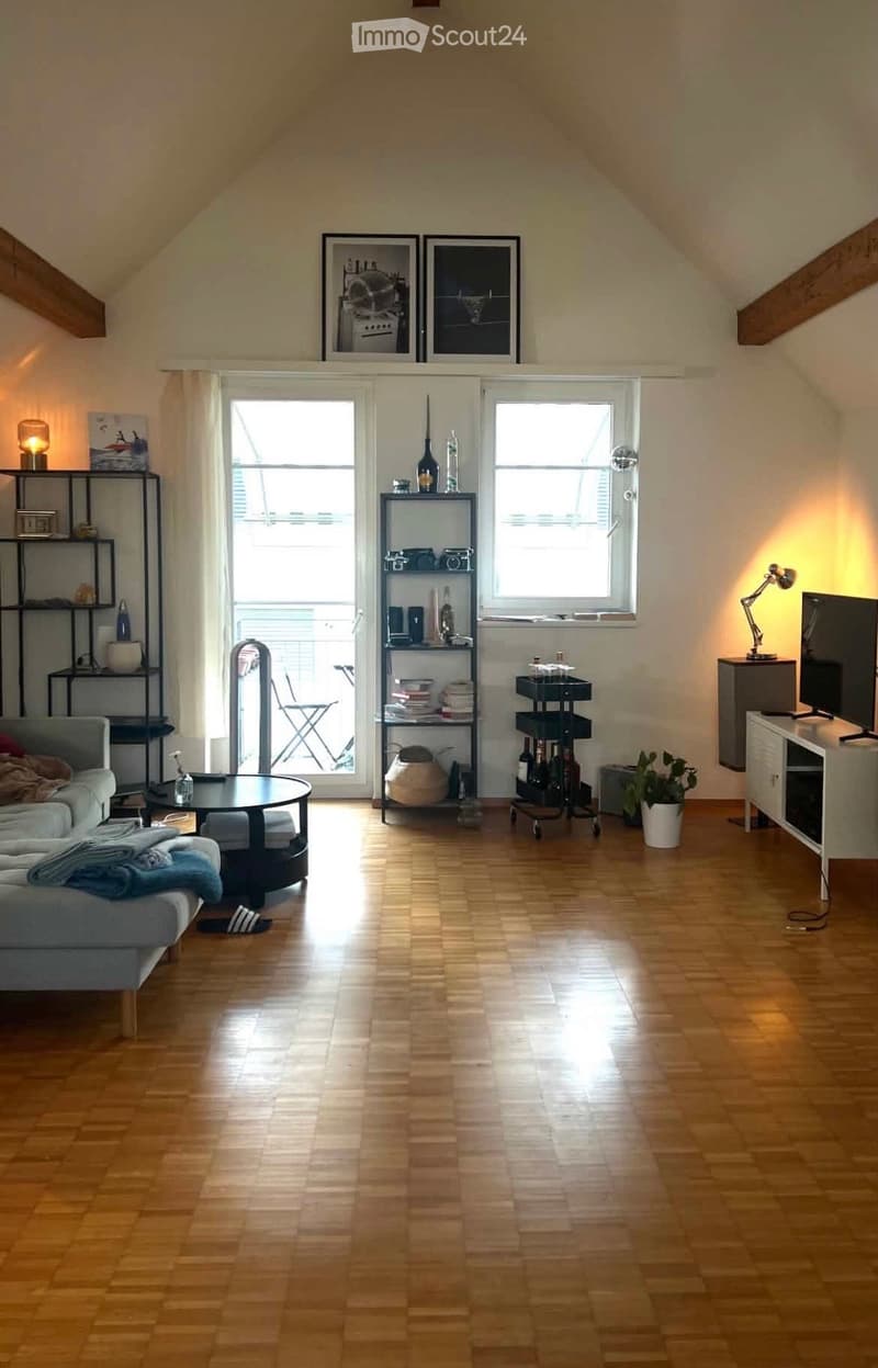 Bright living room with wooden flooring, large windows, and a sliding glass door leading to a balcony. Features a grey sofa, coffee table, shelves with books and decor, and a TV stand with a television.