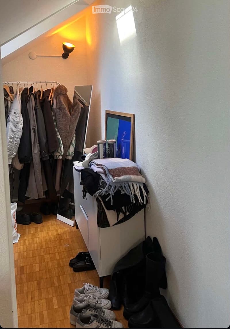 Attic room with wooden flooring, slanted ceiling, and large window. Contains a wardrobe with hanging clothes, a mirror, a picture frame, books, folded blankets, and several pairs of shoes.