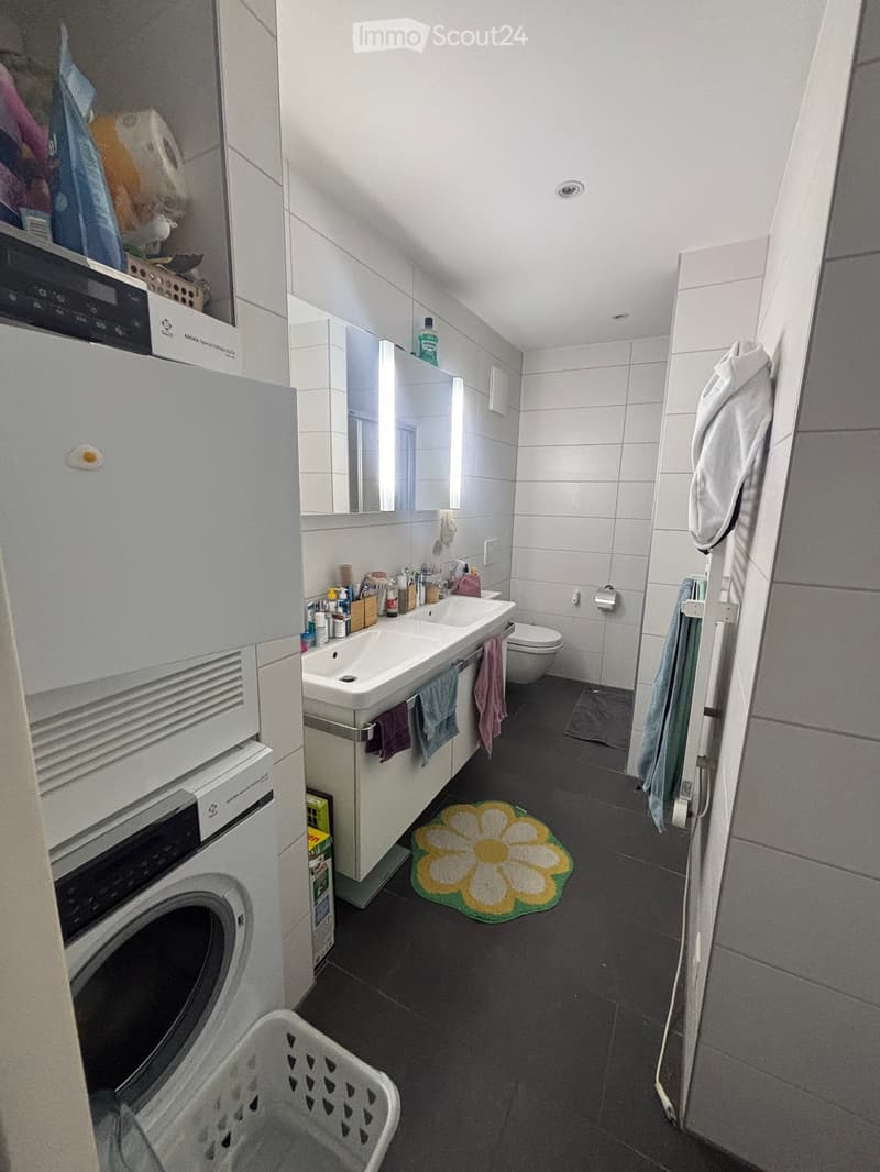 Modern bathroom with white tiles, double sink, toilet, mirror, towels, washing machine, and a flower rug on the floor.