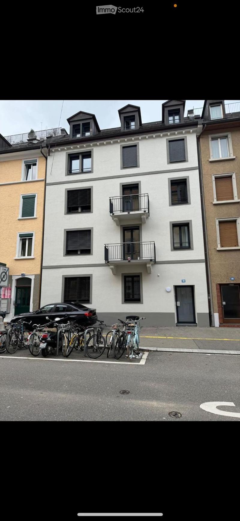 A multi-story residential building with white facade, multiple windows, and balconies. Several bicycles and a car are parked in front.