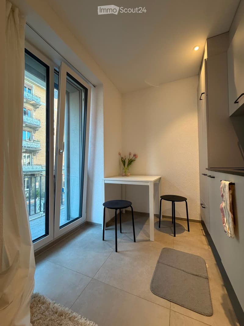 Small, well-lit room with a white table, two black stools, a vase with flowers, a gray rug, and a curtain covering a large window that offers a view of a balcony.