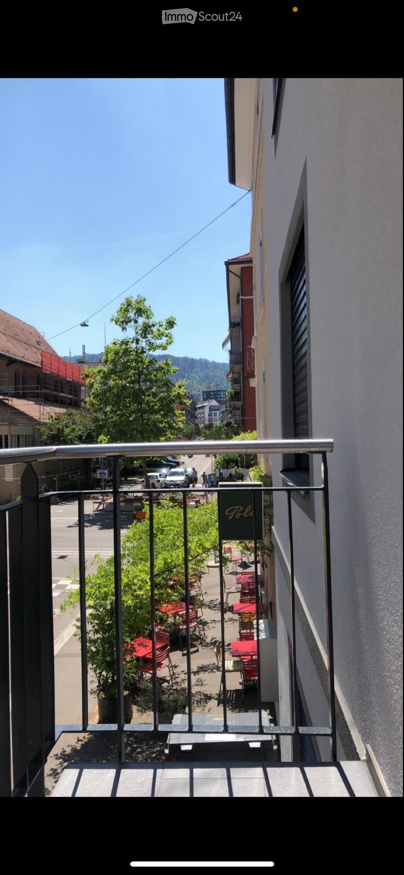 Balcony overlooking a street with outdoor seating, tables, and chairs, surrounded by buildings and trees.