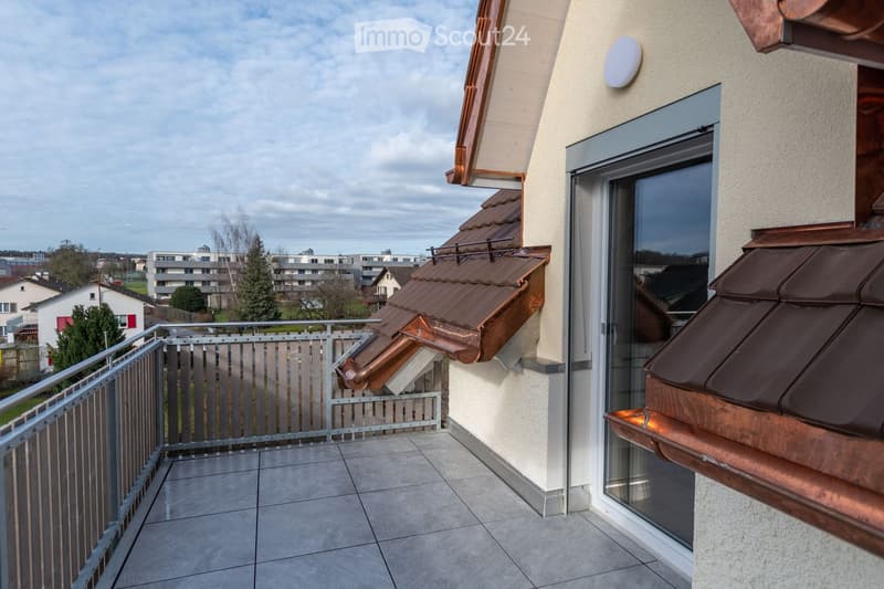 Balcony with tiled floor, metal railing, and copper roof overhangs. The view shows a residential area with buildings and trees.