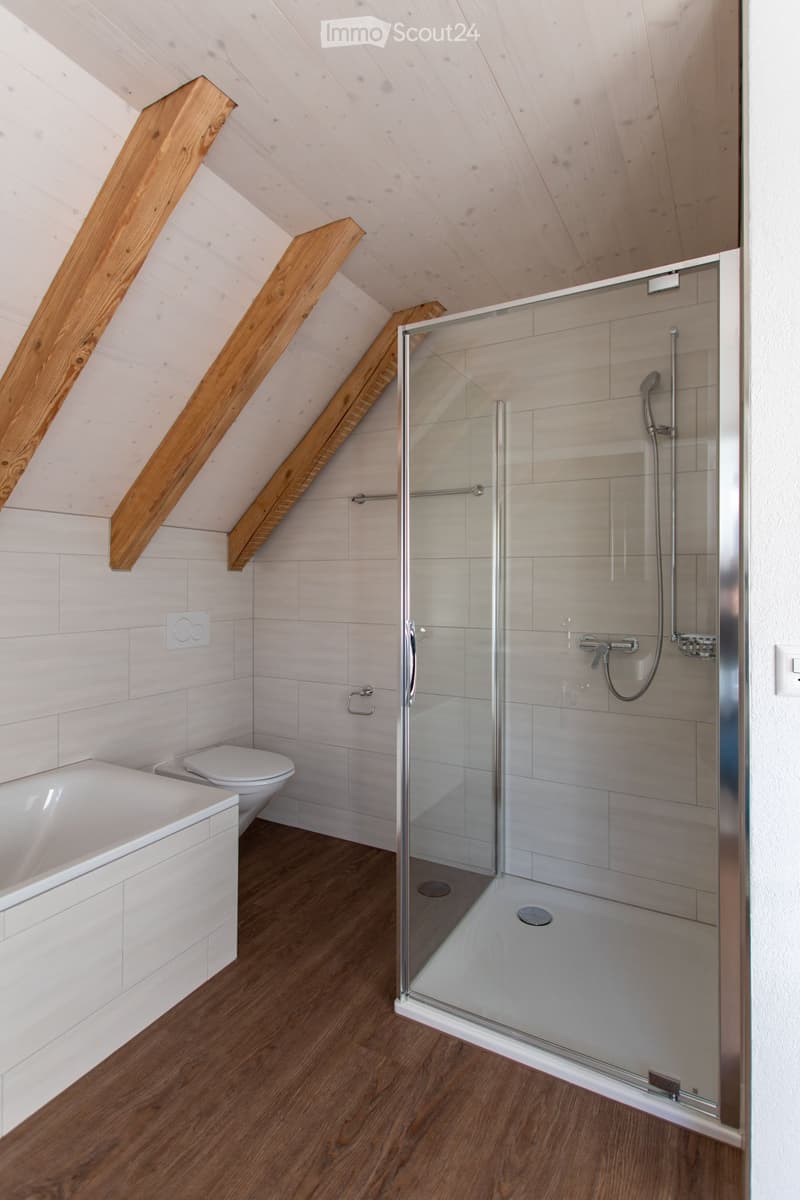 Modern bathroom with a bathtub, toilet, and glass-enclosed shower. The room features light-colored tiles and wooden flooring.