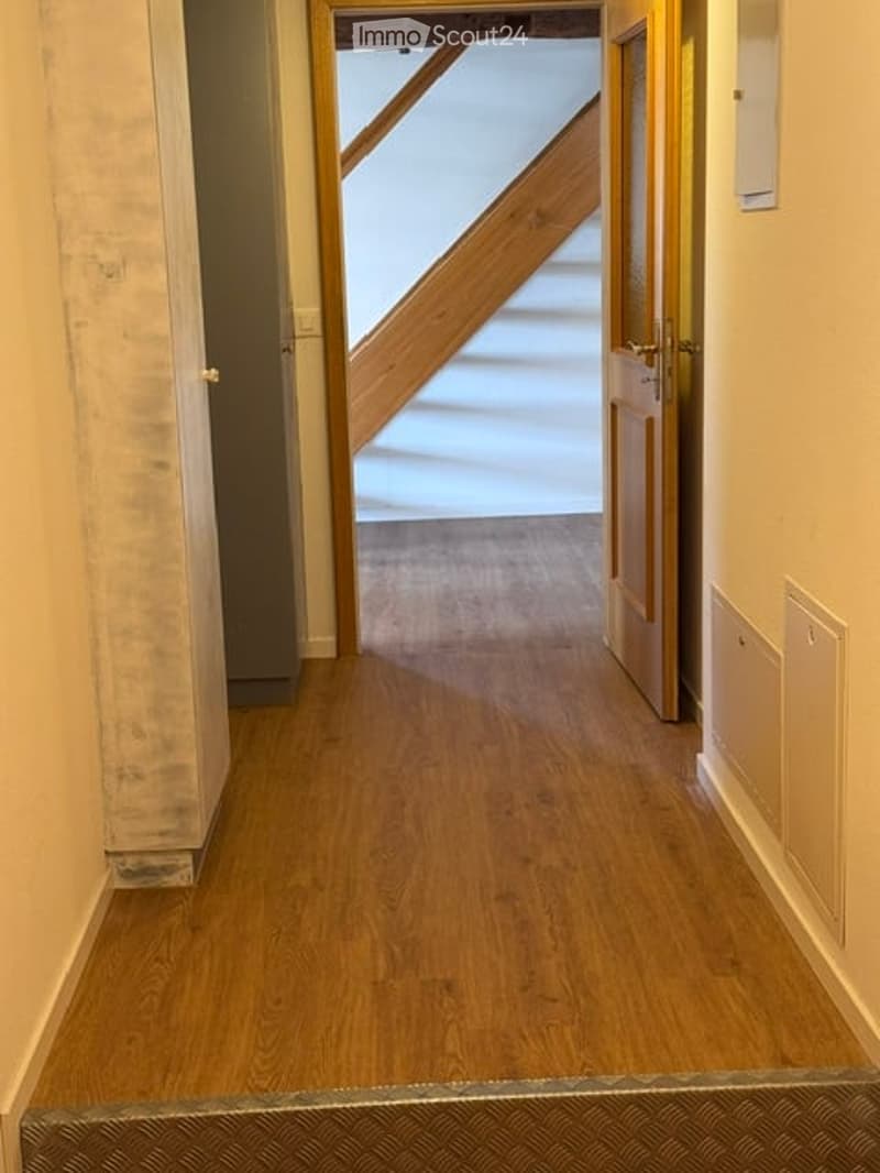 A narrow hallway with wooden flooring, white walls, and a staircase visible at the end.