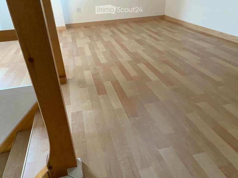 Empty room with light wooden flooring and white walls, showing a corner view with a wooden door frame visible.