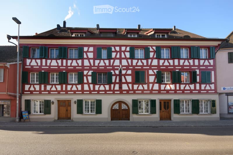 A three-story building with red and white facade, green shutters, multiple windows, and wooden doors.