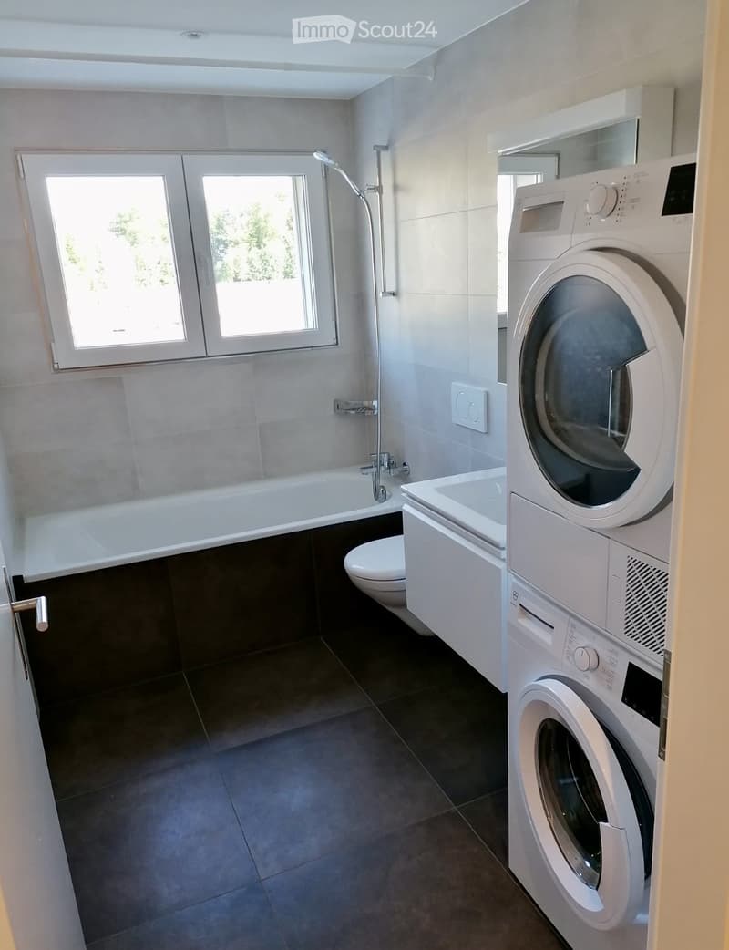 Modern bathroom with a bathtub, toilet, shower, washing machine, and a large window providing natural light.