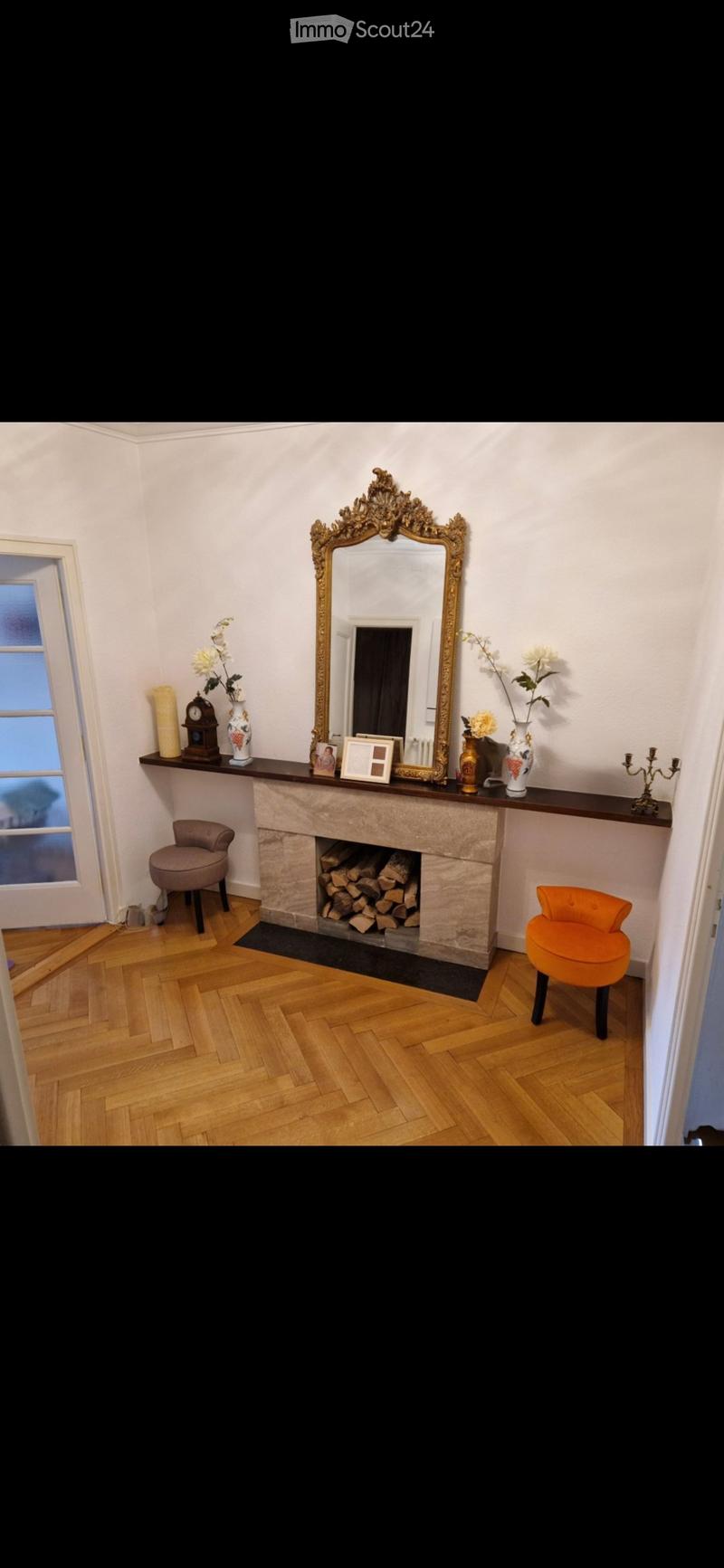 Elegant room with a stone fireplace, two colorful stools, a large ornate mirror, and decorative items on the mantelpiece.
