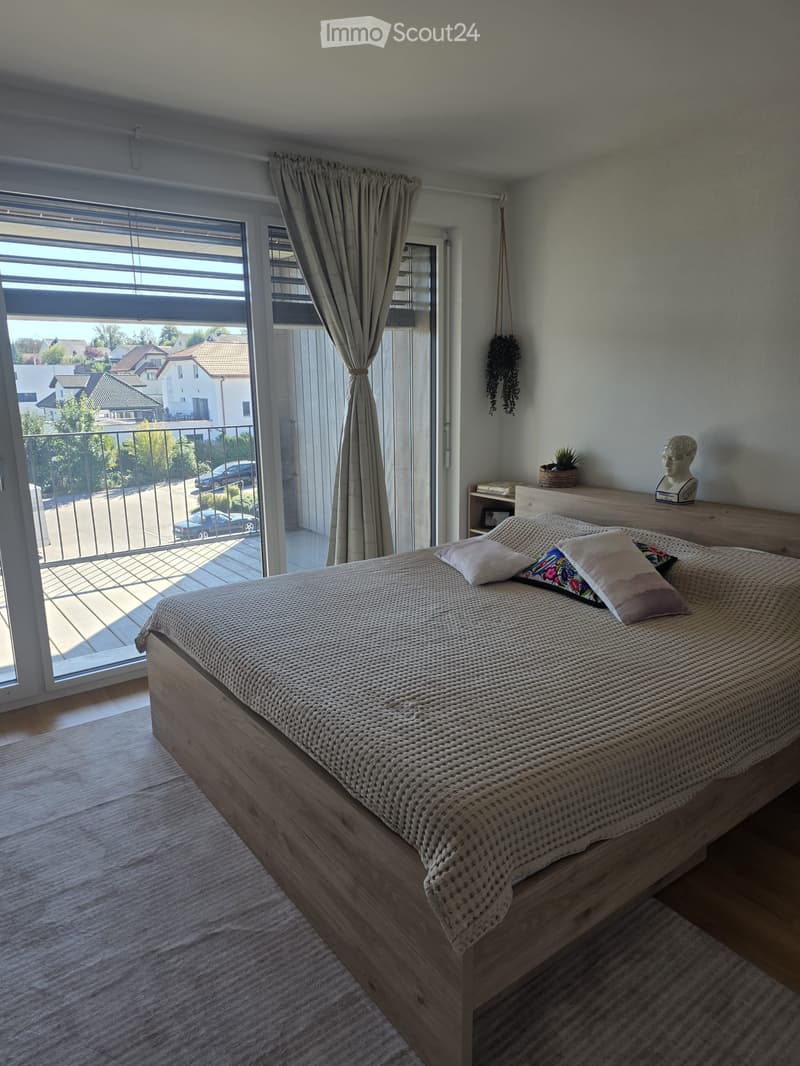Modern bedroom with a wooden bed, decorative pillows, large window with balcony access, and a nightstand with a plant and sculpture.