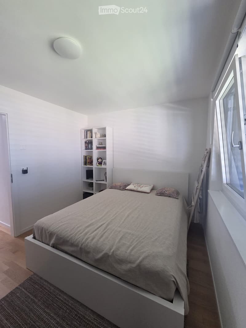 A modern, minimalist bedroom with a white bed, three decorative pillows, a small bookshelf, and a laptop on the shelf. The room features light-colored walls, wooden flooring, and a window with white curtains.