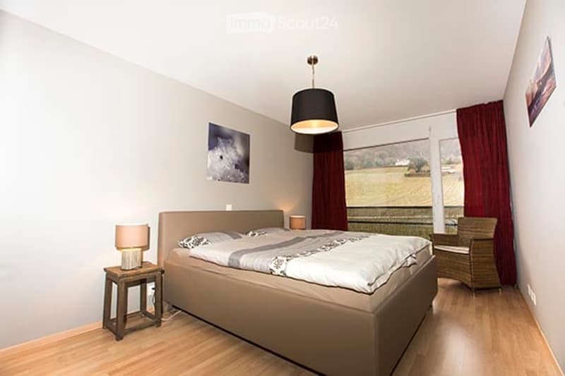Modern bedroom with a large bed, nightstands, lamps, a wicker chair, and artwork on the walls. Large windows with red curtains offer a view of the countryside.