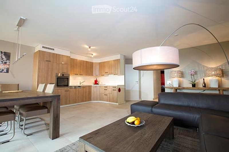 Modern open-plan living and dining area with a sleek black leather sofa, wooden coffee table with fruit bowl, dining table with chairs, and a contemporary kitchen featuring light wood cabinets, stainless steel appliances, and a central island.