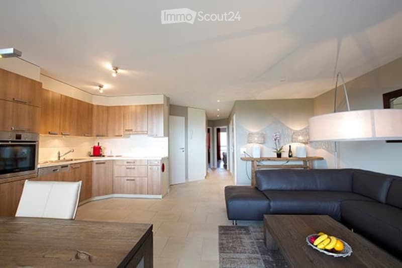Modern open-plan living and dining area with a sleek black sectional sofa, a wooden coffee table with fruit, a dining table with chairs, and a contemporary kitchen featuring light wood cabinets, stainless steel appliances, and a central island.