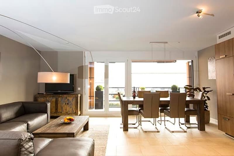 Modern open-plan living and dining area with large windows and balcony access. Features a leather sofa, coffee table, TV, dining table with chairs, and floor lamp.