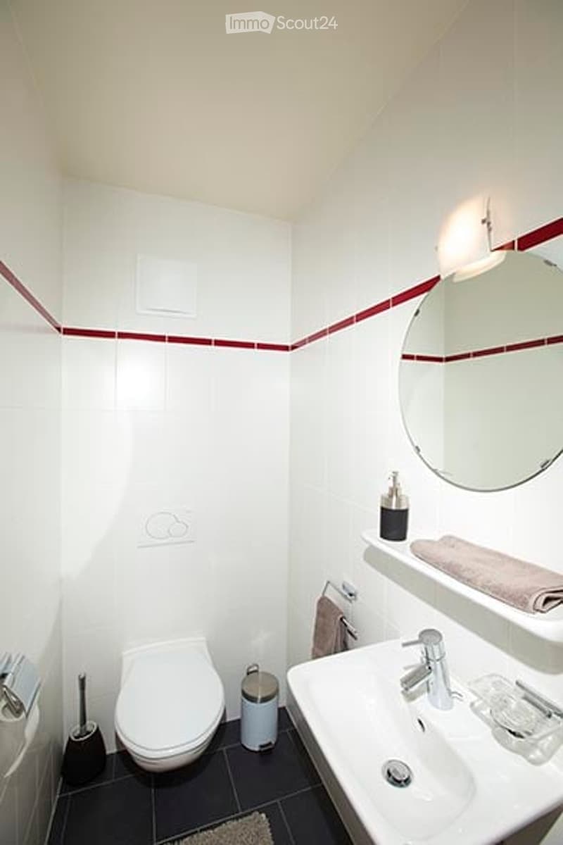 Modern bathroom with white tiles, a toilet, sink, mirror, and towel. Features a red accent border on the wall and a small trash can near the toilet.