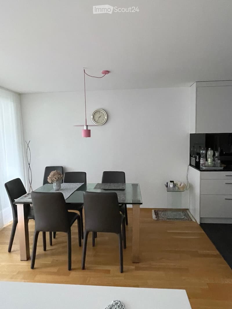 Modern dining area with a glass table, six black chairs, a vase with flowers, a clock on the wall, and a cabinet with drawers.