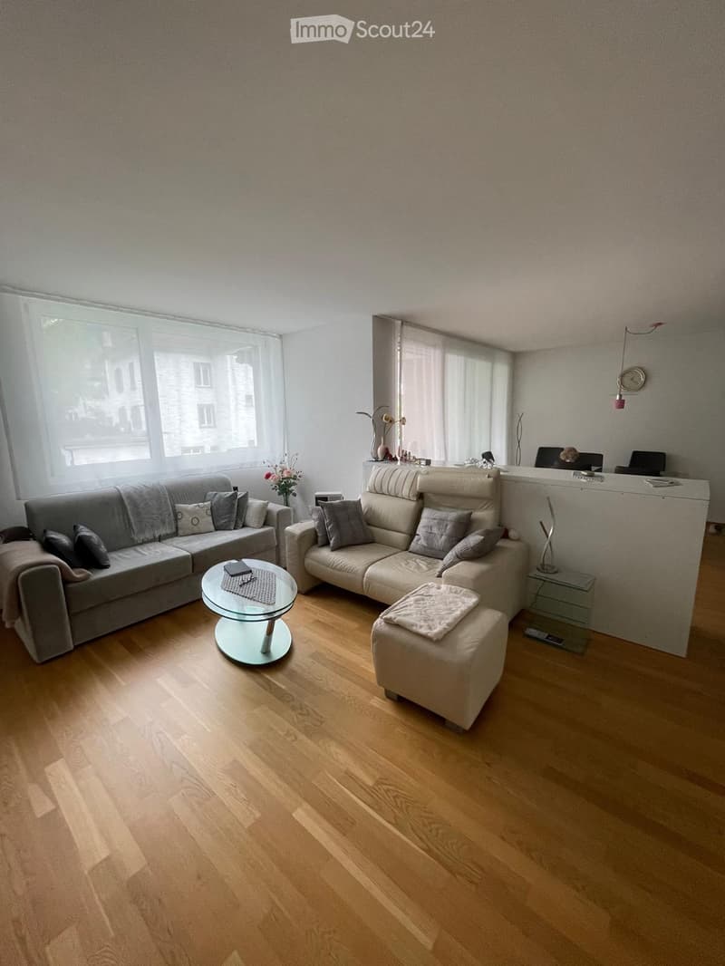 Modern living room with light wooden flooring, two sofas, a coffee table, an ottoman, and a small table with a laptop. Large windows provide natural light.