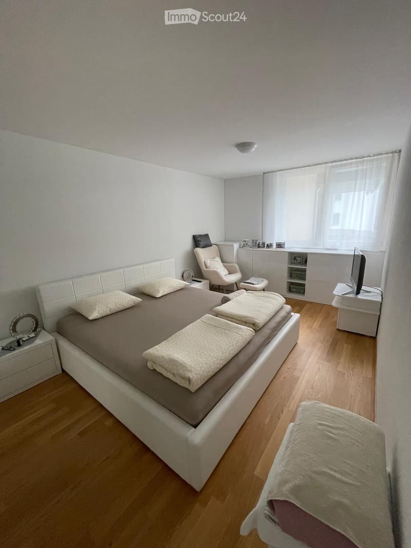 Modern bedroom with a large bed, nightstands, a chair, and a TV screen. Light-colored walls and wooden flooring.