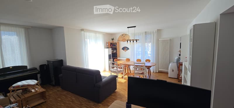 A spacious living and dining area with wooden flooring, a dark blue sofa, a dining table with chairs, a piano, and a fan on the wall.