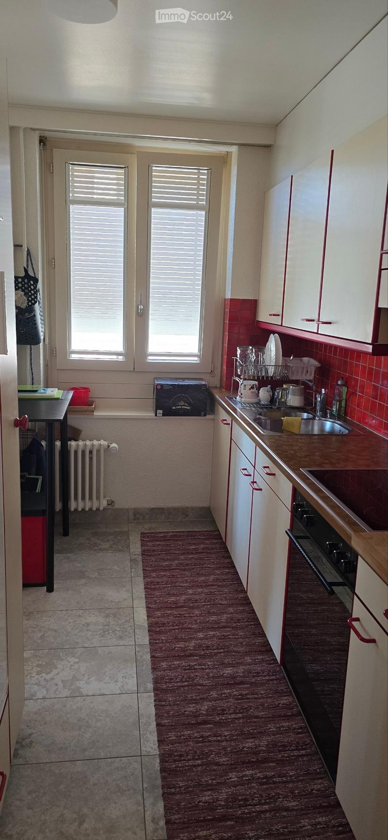 The kitchen features white cabinets with red trim, a wooden countertop, a sink with a faucet, and a dish rack. There is a radiator under the window, a small table with a red container, and a box on the windowsill.