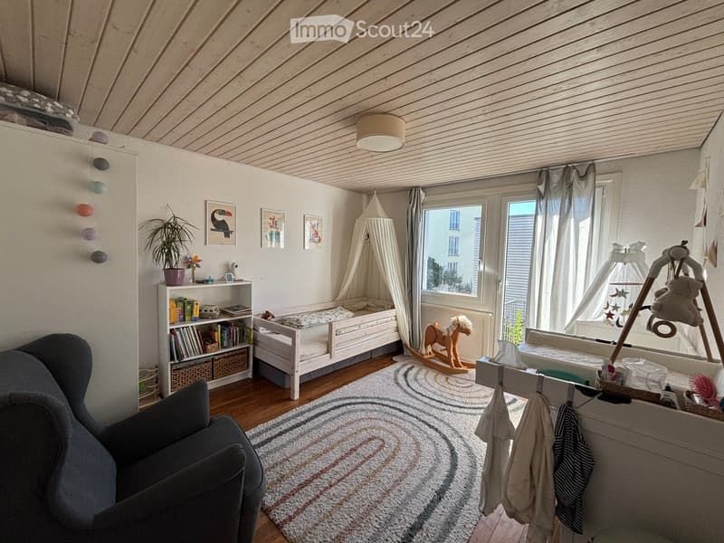 Cozy children's bedroom with a white wooden bed, bookshelf, rocking horse, baby crib, and a soft rug. The room features large windows with sheer curtains and a view of the city.