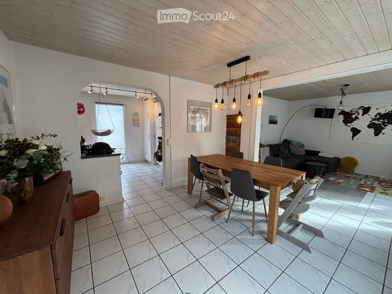 Bright living and dining area with white tiled flooring, wooden dining table with chairs, sofa, baby chair, and decorative elements including a world map and picture frames.