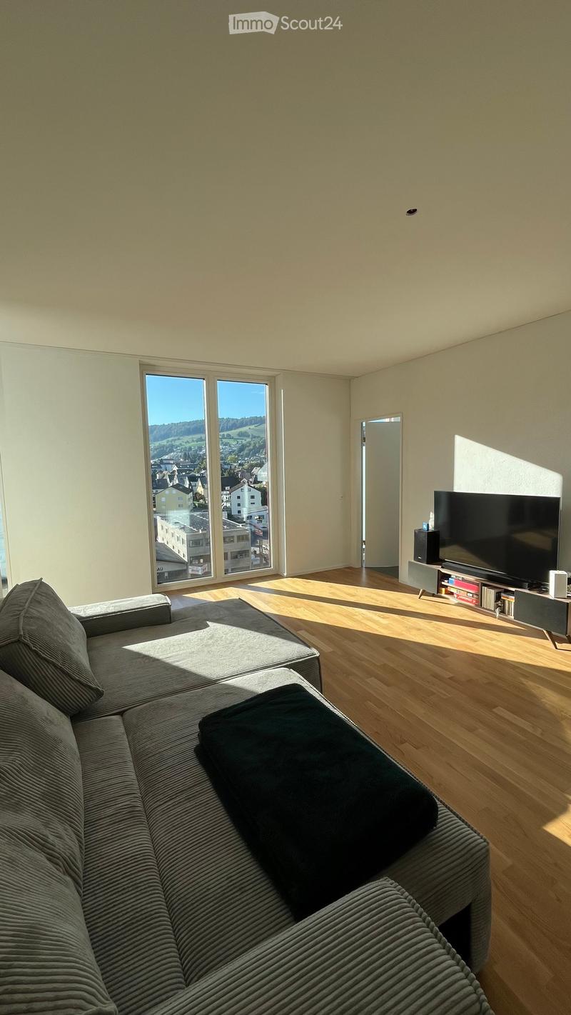 Bright living room with large windows offering a scenic view, featuring a grey sectional sofa, a flat-screen TV on a stand, and a wooden floor.